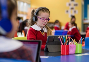 A girl wearing headphones and pink glasses working on a tablet in a colourful classroom