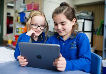 Two girls in blue school uniforms smiling while looking at a tablet together