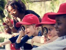 Young children wearing red caps looking through magnifying glasses outdoors with a teacher