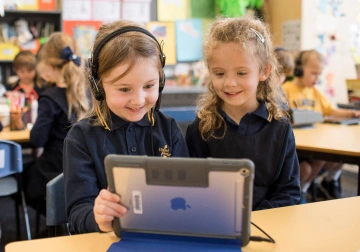 Two young girls smiling while sharing a tablet in a classroom, one wearing headphones